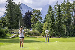 Eine Frau und ein Mann spielen Golf auf einem grünen Platz, umgeben von hohen Bäumen, mit Bergen im Hintergrund. Die Szene ist friedlich und sonnig.
