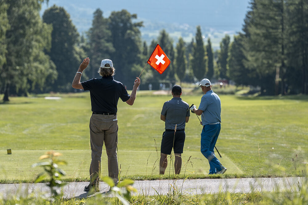 Drei Golfer stehen auf einem üppigen grünen Platz und bewundern einen Schlag. Bäume säumen den Hintergrund unter einem klaren blauen Himmel, der ein Gefühl von Muße und Konzentration hervorruft.