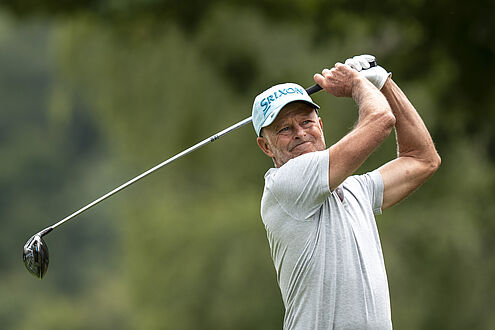 Jean-Francois Remésy in hellblauer Mütze und weißem Hemd schwingt konzentriert und gelassen den Schläger. Das üppige Grün im Hintergrund erinnert an einen ruhigen Golfplatz.