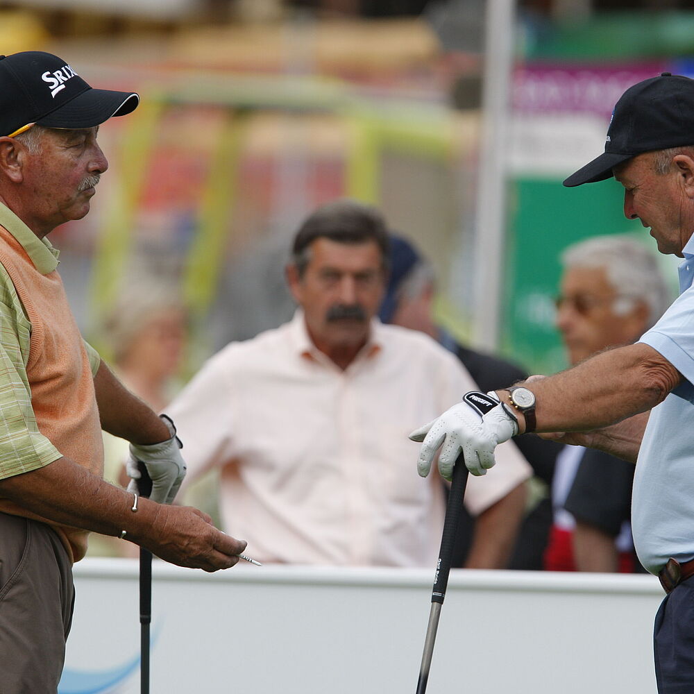 Zwei ältere Männer stehen sich auf einem Golfplatz gegenüber, jeder hält einen Golfschläger. Sie scheinen in ein Gespräch vertieft zu sein, mit einem konzentrierten Ausdruck.
