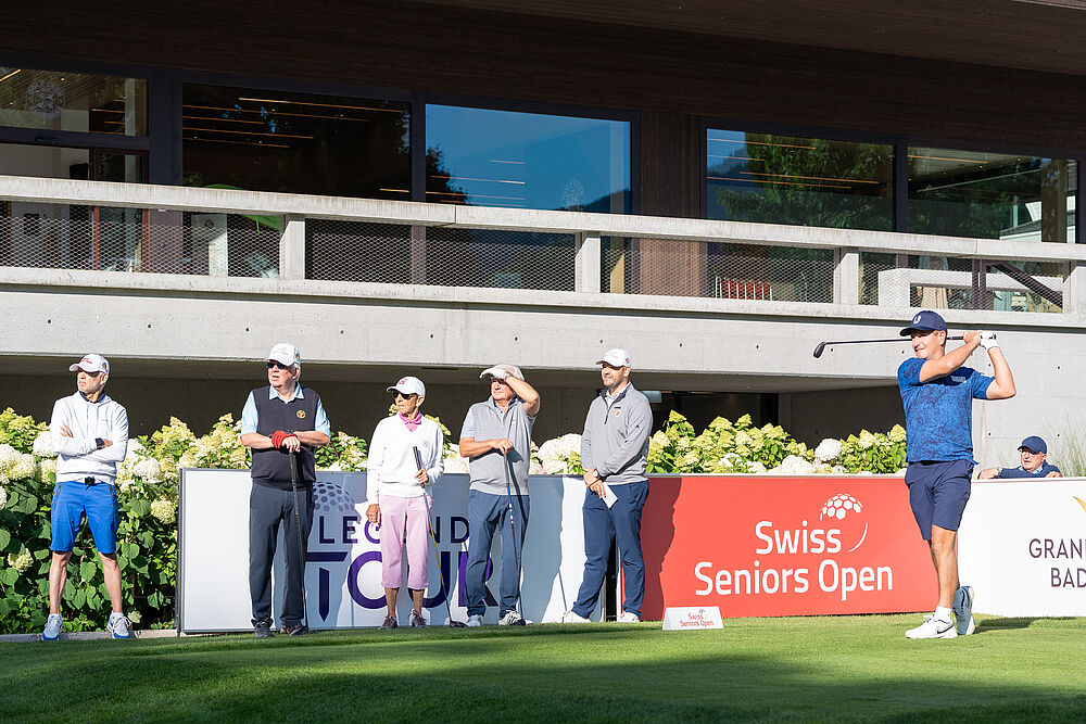 Eine Gruppe von sechs Personen in Golfkleidung steht auf einem Golfplatz, einige halten Schläger. Sie scheinen konzentriert zu sein. Im Hintergrund sind Banner und Grünpflanzen zu sehen.