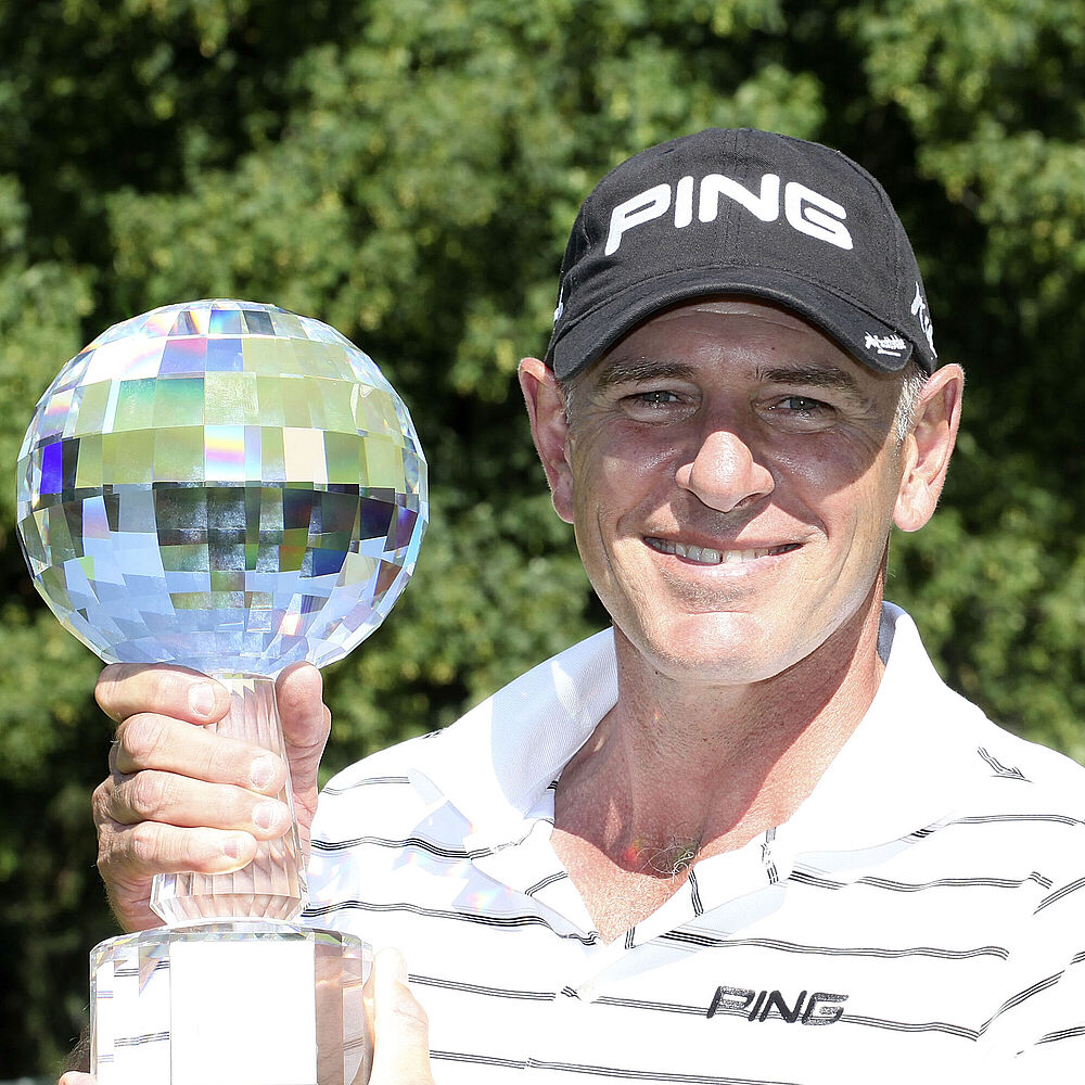 Peter Fowler in einem gestreiften Hemd und einer PING-Mütze lächelt, während er eine glänzende, kugelförmige Trophäe hält. Helle Umgebung im Freien mit grünen Bäumen im Hintergrund, die den Triumph vermitteln.