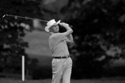 Ein Herr der nach dem Golf Abschlag seinem Ball nachschaut und den Schläger noch über den Schultern hält. Er trägt einen weissen Cowboy Hut. Das Bild ist in schwarz weiss. 