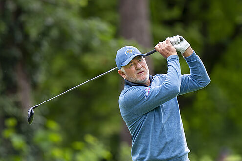 Peter Baker in einer blau gemusterten Jacke und einem Cap schwingt einen Schläger inmitten von Grün und vermittelt so Konzentration und Präzision während eines ruhigen Golfspiels.