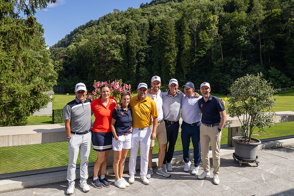 Eine Gruppe von acht Leuten steht nebeneinander Arm in Arm. Die beiden Frauen tragen kein Cap. Sie sind farbig gekleidet und im hintergrund ist der Golfplatz und rechts ein Wald zu sehen.
