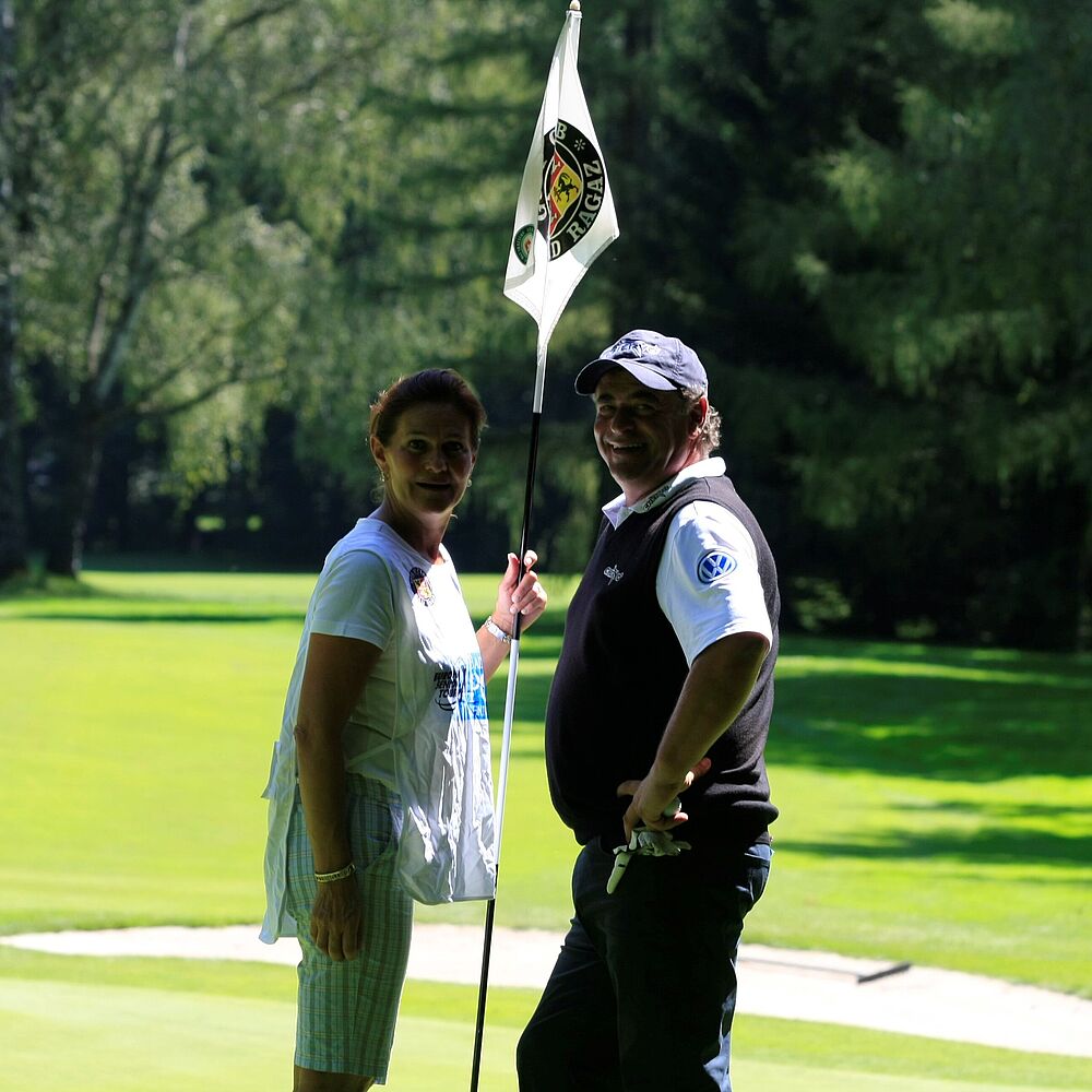 Eine Frau und Costantino Rocca stehen auf einem sonnigen Golfplatz in der Nähe eines Lochs mit einer Fahne. Sie wirken entspannt, umgeben von saftig grünem Gras und hohen Bäumen.