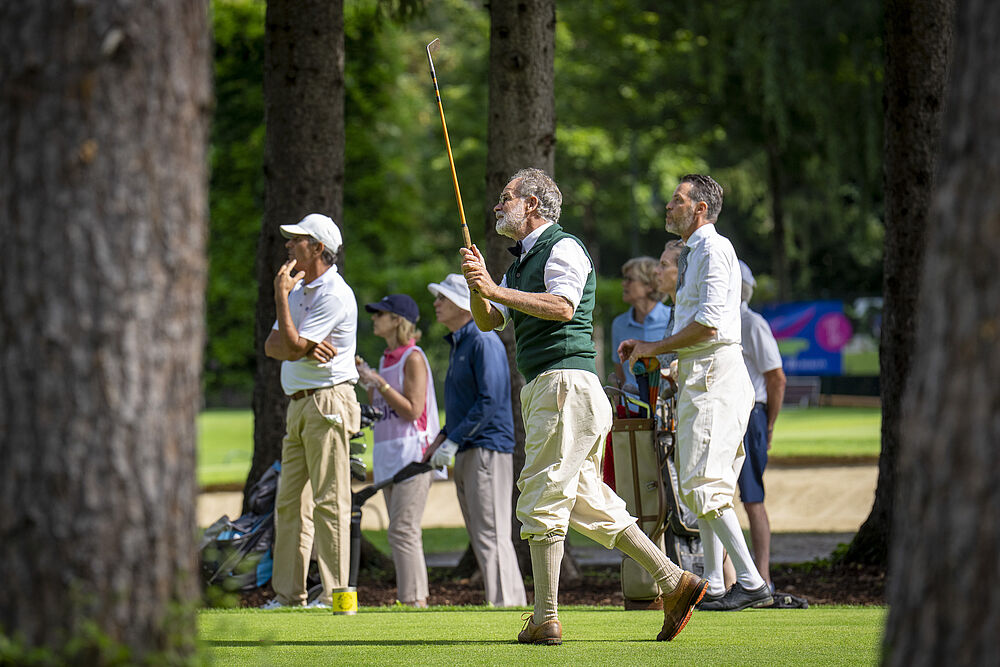 Eine Gruppe von Menschen spielt Golf. Zwei Ein Mann schwingt den Schläger in der Luft und trägt eine grüne Weste. Andere schauen zu, umgeben von hohen Bäumen.