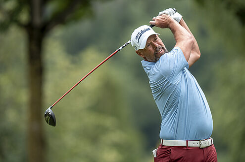 Mikael Lundberg in einem blauen Hemd und einer roten Hose schwingt einen Schläger auf einem Golfplatz. Bäume und Grünflächen verschwimmen im Hintergrund und vermitteln Konzentration und Entschlossenheit.