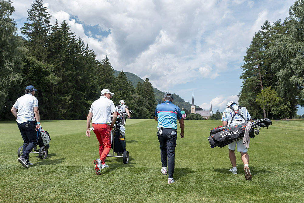 Eine Gruppe von Golfern und Caddies geht über einen grünen Golfplatz unter einem wolkenverhangenen blauen Himmel, umgeben von hohen Bäumen und fernen Bergen.