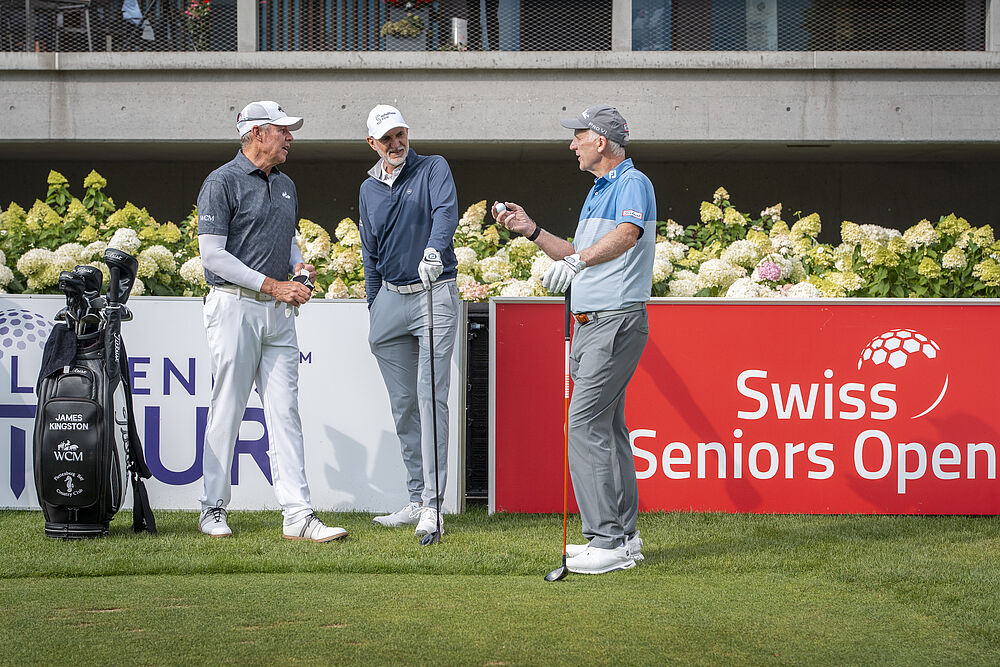 Drei Golfer in Golfkleidung stehen auf einem Golfplatz, unterhalten sich in der Nähe eines roten „Swiss Seniors Open“-Schildes, die Golfschläger an der Seite.