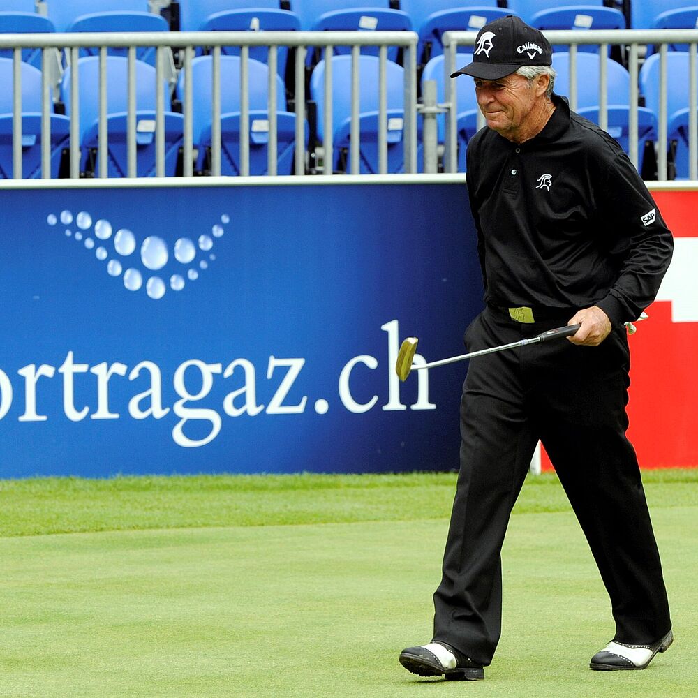 Bob Verwey in schwarzer Kleidung geht auf dem Grün und hält einen Putter. Blaue Stände und Werbebanner im Hintergrund. Konzentrierte und ruhige Szene.