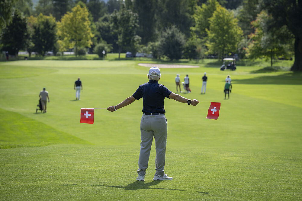 Eine Person steht mit ausgestreckten Armen auf einem Golfplatz und beobachtet die Spieler in der Ferne. Fahnen markieren das saftig grüne Feld an einem sonnigen Tag.