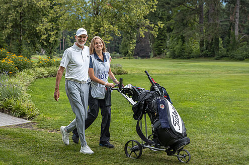 Golfprofi André Bosser und seine Tochter laufen über den Golfplatz. Sie schiebt den Golfbag vor sich her. Beide sind hell gekleidet und im Hintergrund ist viel grün.