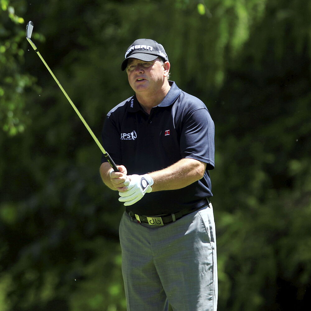 Ian Woosnam in einem marineblauen Hemd und einer Mütze hält einen Schläger und steht auf dem sonnigen Golfplatz. Er scheint konzentriert zu sein, mit dichtem Grün im Hintergrund.