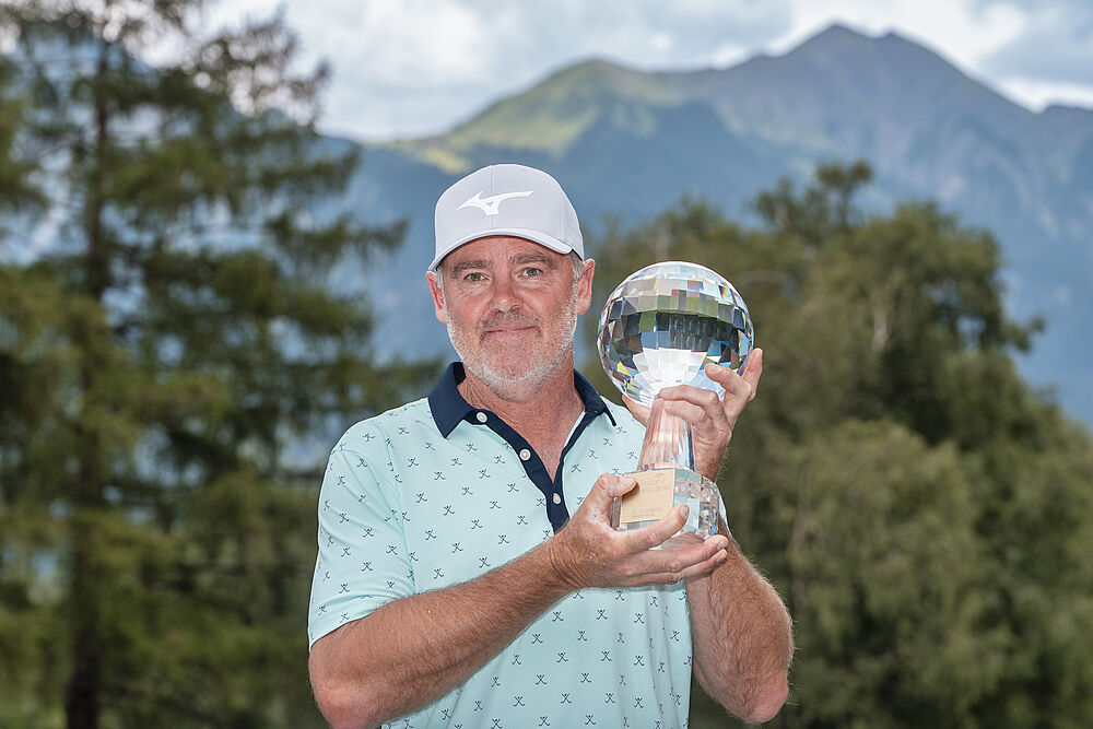 Mark Brown hält den Kristallpokal in die Höhe. Trägt ein helles Shirt und ein weisses Cap. Im Hintergrund sind Bäume zu sehen.