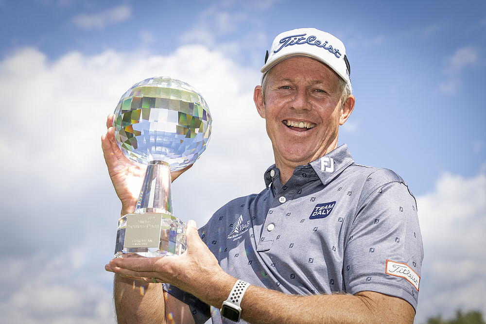 James Kingston mit Golfmütze und gemustertem Hemd hält stolz die Glastrophäe vor einem strahlend blauen Himmel mit vereinzelten Wolken.