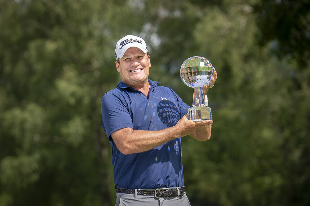 Jarmo Sandelin, der Sieger des Swiss Seniors Open 2024 hält den Glaspokal in die Lüfte. Er trägt ein blaues Hemd und ein weisses Cap und lächelt, während er auf dem Golfplatz die Trophäe hält. Grüne Bäume bilden einen unscharfen Hintergrund.