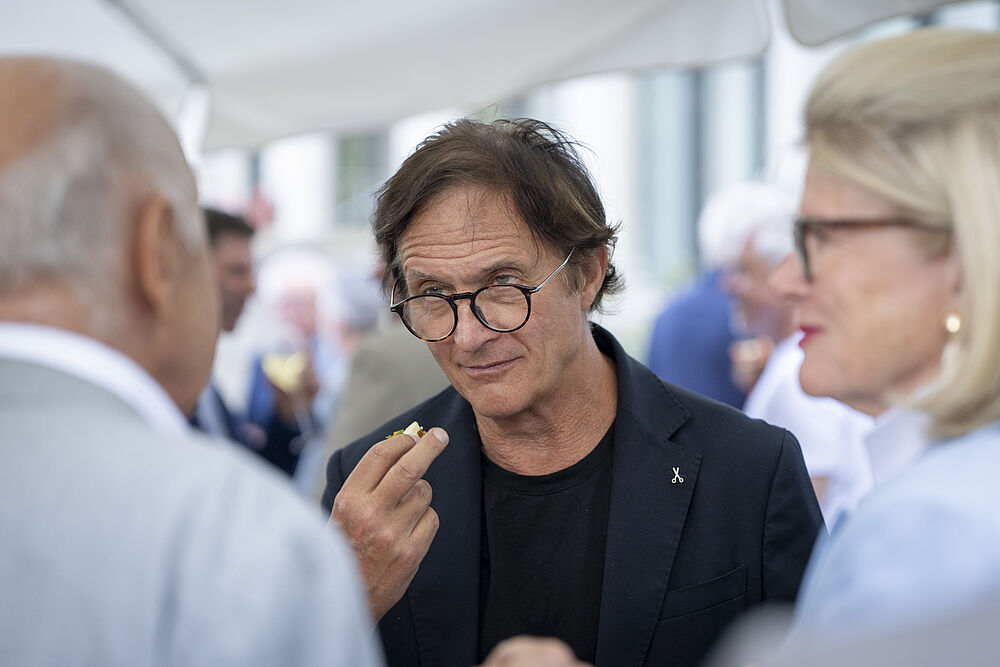 Arno del Curto mit Brille, der einen Snachk in der Hand hält, unterhält sich mit zwei Personen unter einem weißen Regenschirm. Sie sind am Gala Abend anlässlich des ProAm's im Aussenbereich des Kursaals. Es herrscht eine gesellige und lebhafte Atmosphäre.
