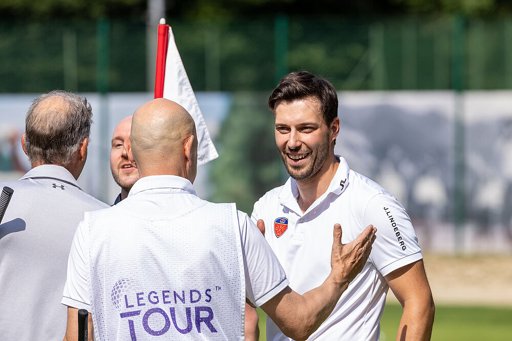 Drei Männer in weißen Hemden stehen auf einem sonnigen Golfplatz und unterhalten sich. Auf einem Hemd steht „Legends Tour“. Die Atmosphäre ist entspannt und freundlich.