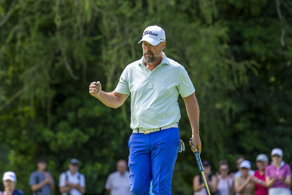 Ein Golfer feiert einen erfolgreichen Schlag mit erhobener Faust vor einer Kulisse aus unscharfen Zuschauern und üppigem Grün, die Triumph und Aufregung vermittelt.