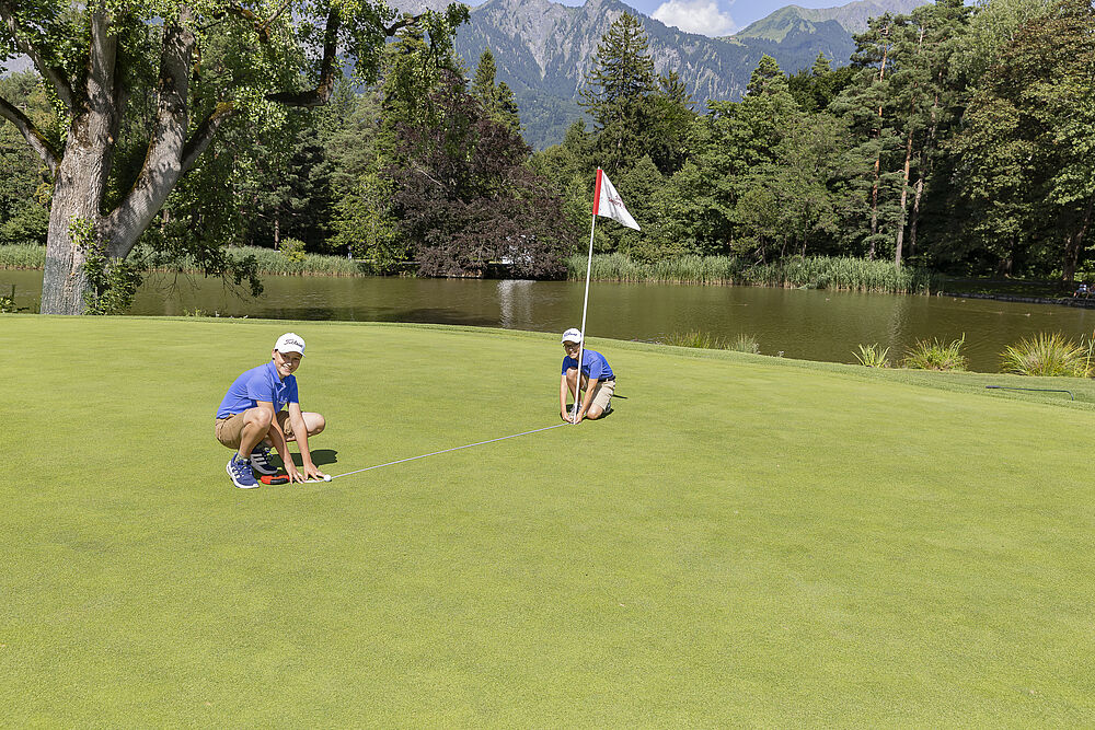 Zwei Golfer in blauen Hemden und weißen Mützen hocken auf einem üppig grünen Golfplatz in der Nähe eines Teichs. Im Hintergrund sind Bäume und Berge unter einem klaren Himmel zu sehen.
