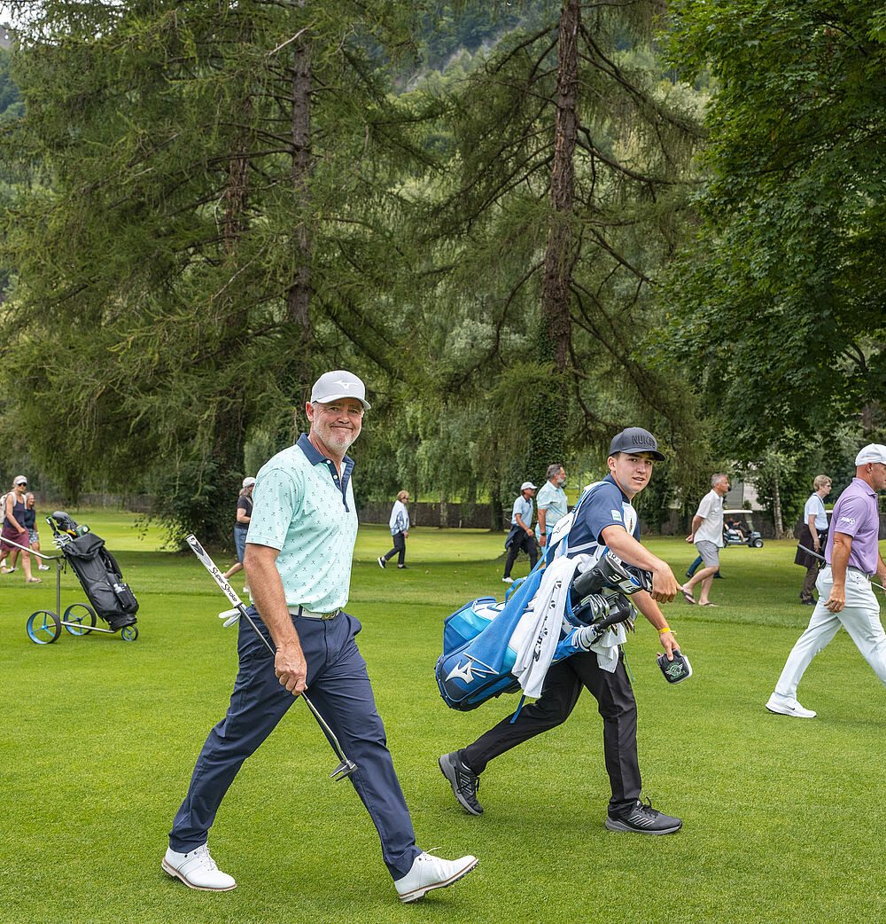 Sieger Mark Brown und sein Caddy laufen über den Golfplatz. Er ist in blau gekleidet mit weissem Cap. Hinter ihnen her laufen einige Zuschauer. Er lächelt in die Kamera und wirkt fröhlich.