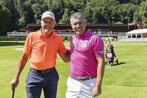 Bernhard Russi und Adilson da Silva auf einem Golfplatz; einer im orangefarbenen Hemd, der andere in Rosa. Heller sonniger Tag, Clubhaus im Hintergrund, üppiges grünes Fairway.