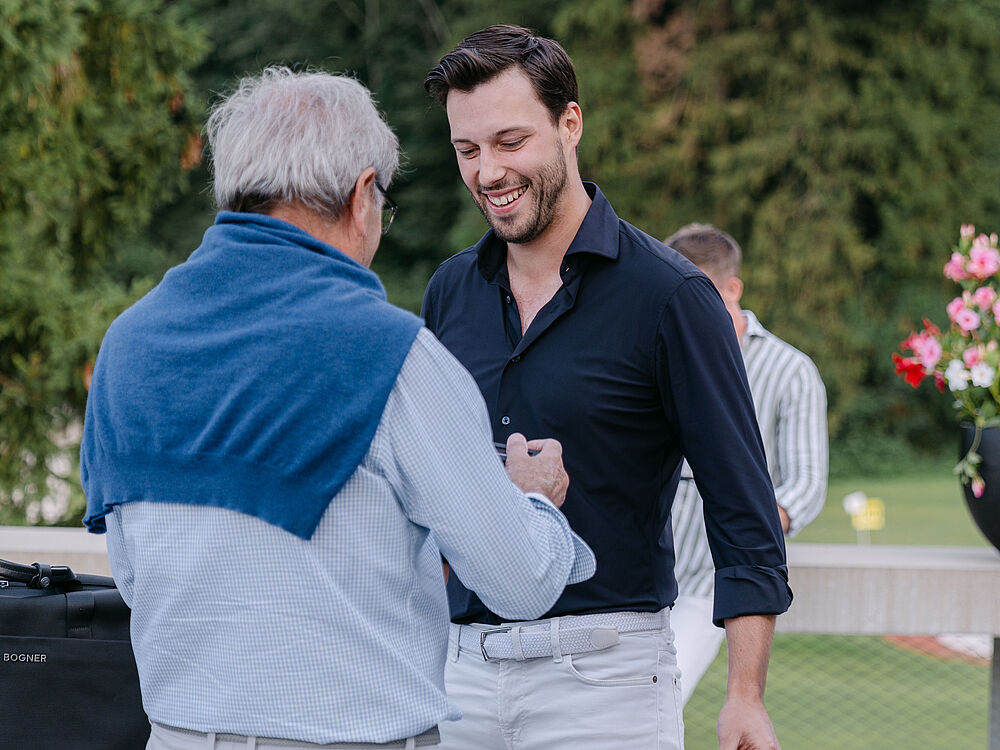 Ein Mann mit blauem Pulli über den Schultern übergibt ein Geschenk an den anderen Mann der ein dunkles Hemd trägt. Dieser lächelt und freut sich über die Geste. Sie schütteln sich die Hand.