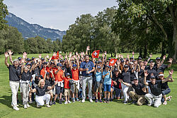 Eine Gruppe von Menschen, die Helfer des Swiss Seniors Open, posiert fröhlich auf dem grünen Golfplatz, umgeben von Bäumen und Bergen, und viele heben feierlich die Arme. In der Mitte steht Jarmo Sandelin, der Sieger von 2024 mit seinem Glaspokal.