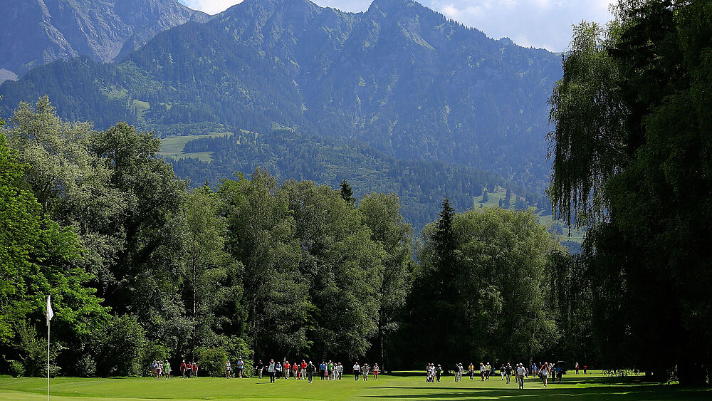 Ein malerischer Golfplatz mit Spielern in farbenfroher Kleidung auf einem grünen Fairway, umgeben von üppigen Bäumen und majestätischen Bergen unter einem teilweise bewölkten Himmel.