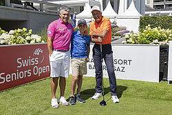 Adilson da Silva links, in der Mitte sein Sohn und rechts Bernhard Russi stehen auf dem Golfplatz mit einem Turnierschild im Hintergrund. Sie lächeln, sind in bunte Golfkleidung gekleidet und vermitteln eine fröhliche Stimmung.