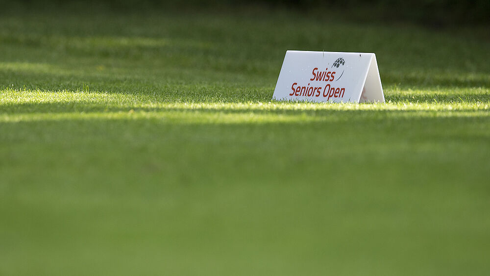 Ein Schild auf einem grünen Golfplatz zeigt in roter Schrift „Swiss Seniors Open“. Das Sonnenlicht erzeugt Muster auf dem Gras und vermittelt eine ruhige, konzentrierte Atmosphäre.