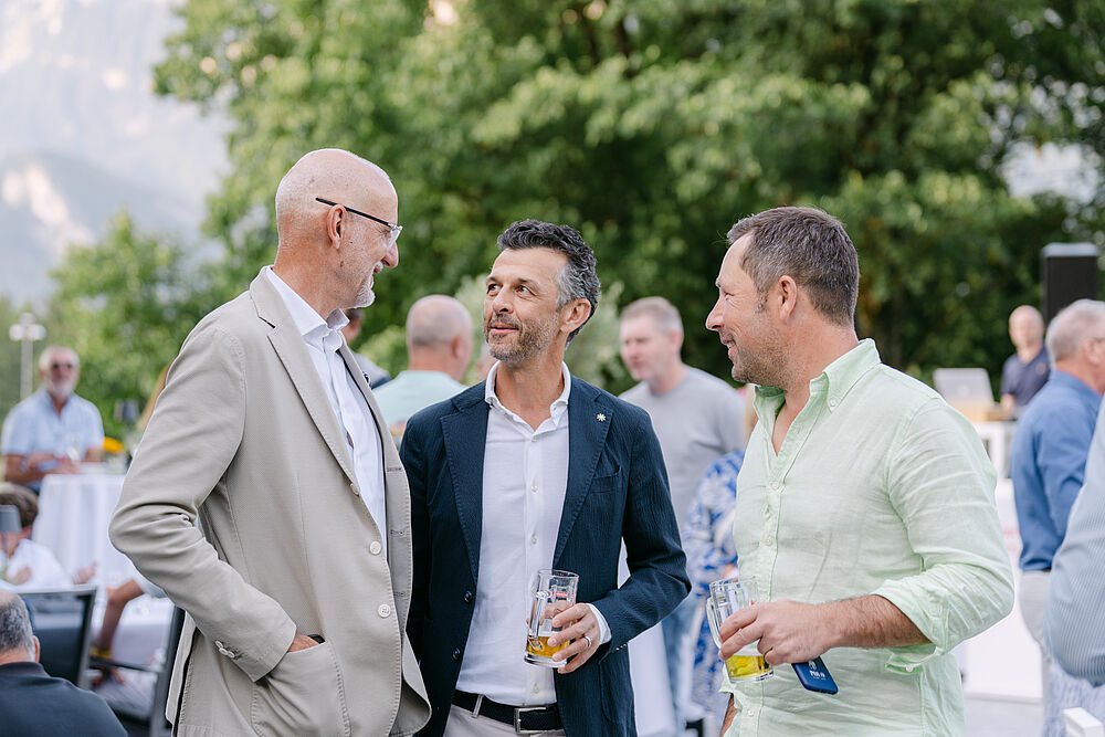 Drei Männer in festlicher Kleidung unterhalten sich. Der links trägt baige. Der in der Mitte trägt ein weisses Hemd mit dunklem Jacket und rechts trägt der Mann ein Hemd in leichtem grün. Im Hintergrund sin Bäume zu sehen.