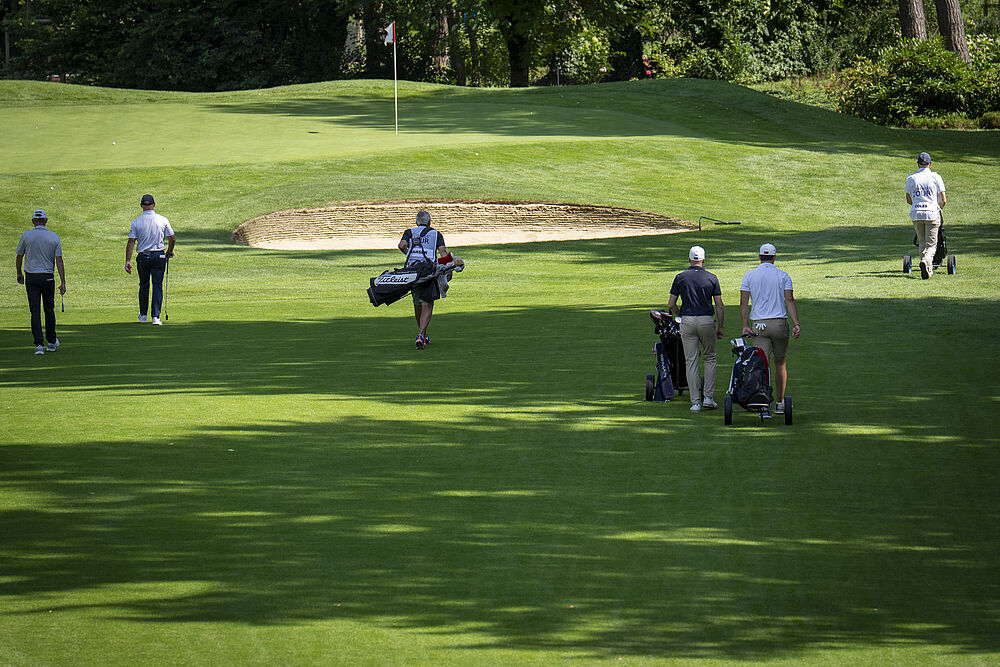 Golfer und Caddies nähern sich auf einem sonnenbeschienenen Grün einem Bunker, umgeben von üppigen Bäumen. Die Szene vermittelt Konzentration und Vorfreude in einer heiteren Umgebung.