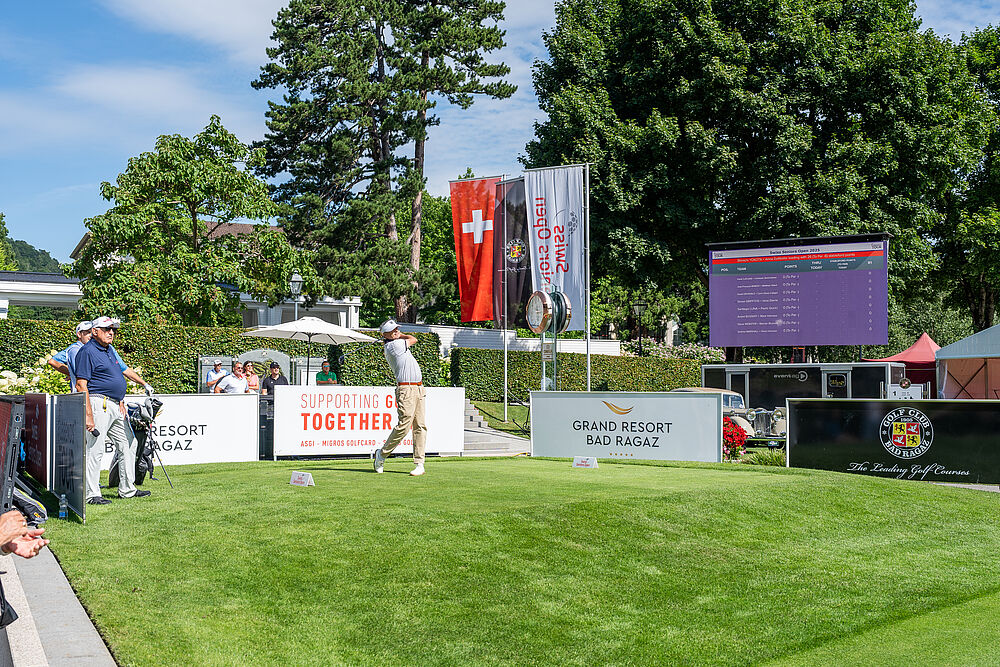 Ein Golfer steht beim Abschlag auf saftig grünem Rasen und hat soeben den Ball gespielt. Hinter ihm sind Werbebanden und drei Fahnen zu sehen. Rechts davon ein grosser Bildschirm der das Leaderboard zeigt.  