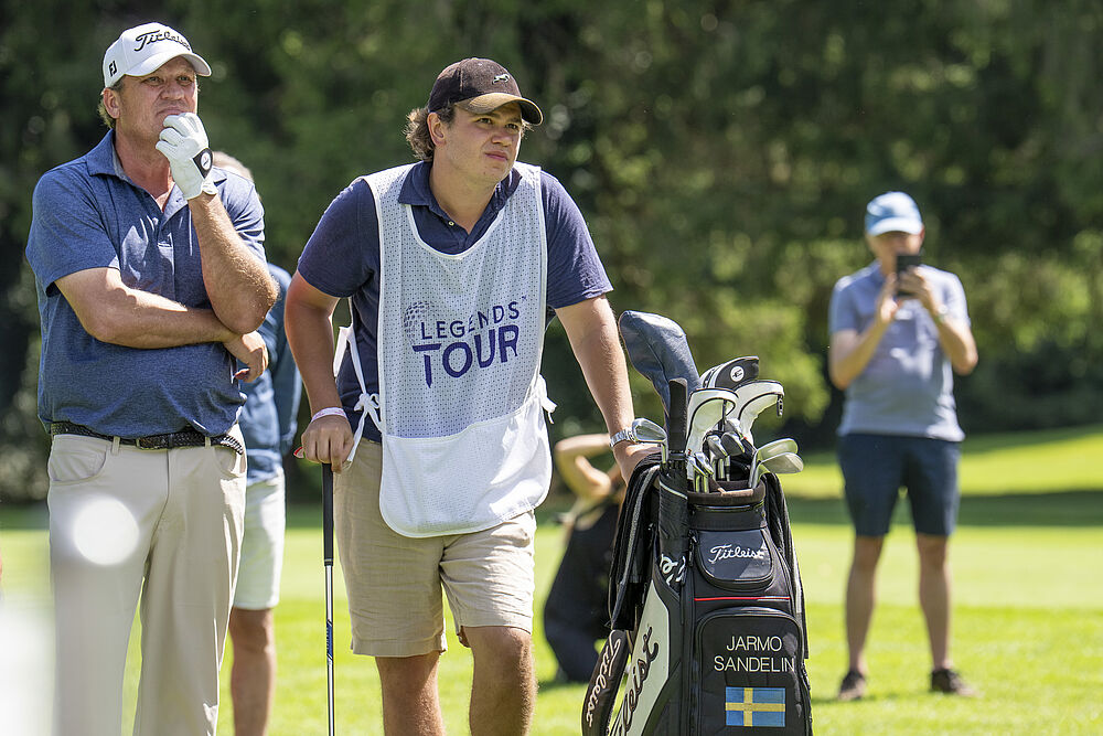 Zwei Personen stehen auf einem Golfplatz, die eine in einer weißen Caddie-Weste, die andere in einem blauen Hemd, beide konzentriert. Grünes Gras und Bäume im Hintergrund.