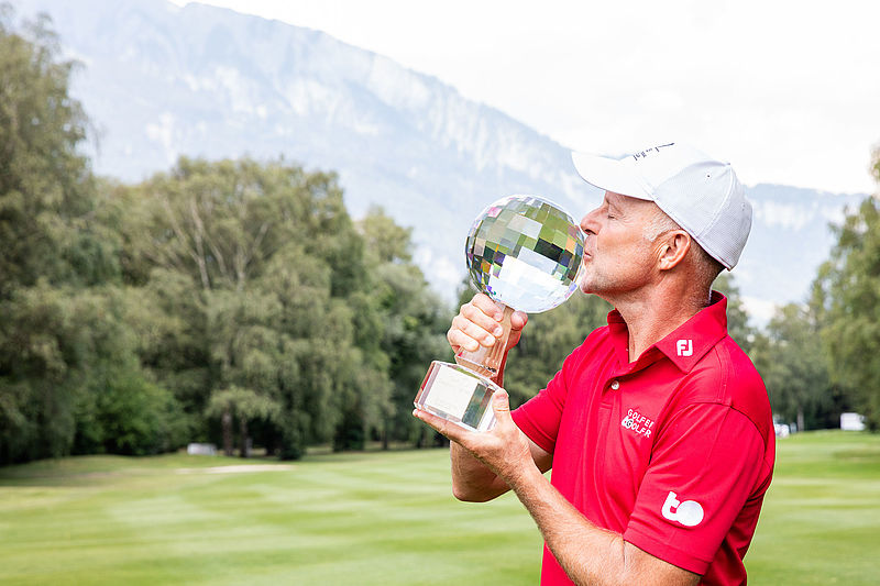 Jean-François Remésy in rotem Hemd und weißer Mütze küsst den Glaspokal auf dem Golfplatz, mit fernen Bergen und Bäumen im Hintergrund, die Triumph ausstrahlen.