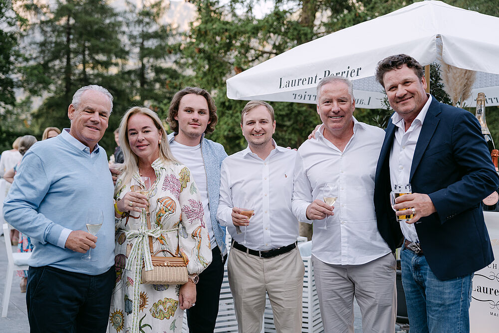 Eine Gruppe von sechs Personen. Alle in festlicher Kleidung und mit Weingläser in der Hand. Rechts im Hintergrund ist ein weisser Sonnenschirm zu sehen.
