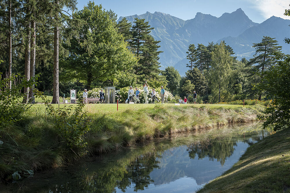 Eine ruhige Golfrunde im Freien in der Nähe eines spiegelnden Teichs mit üppigen Bäumen und hoch aufragenden Bergen im Hintergrund unter einem klaren blauen Himmel. Eine ruhige Umgebung.