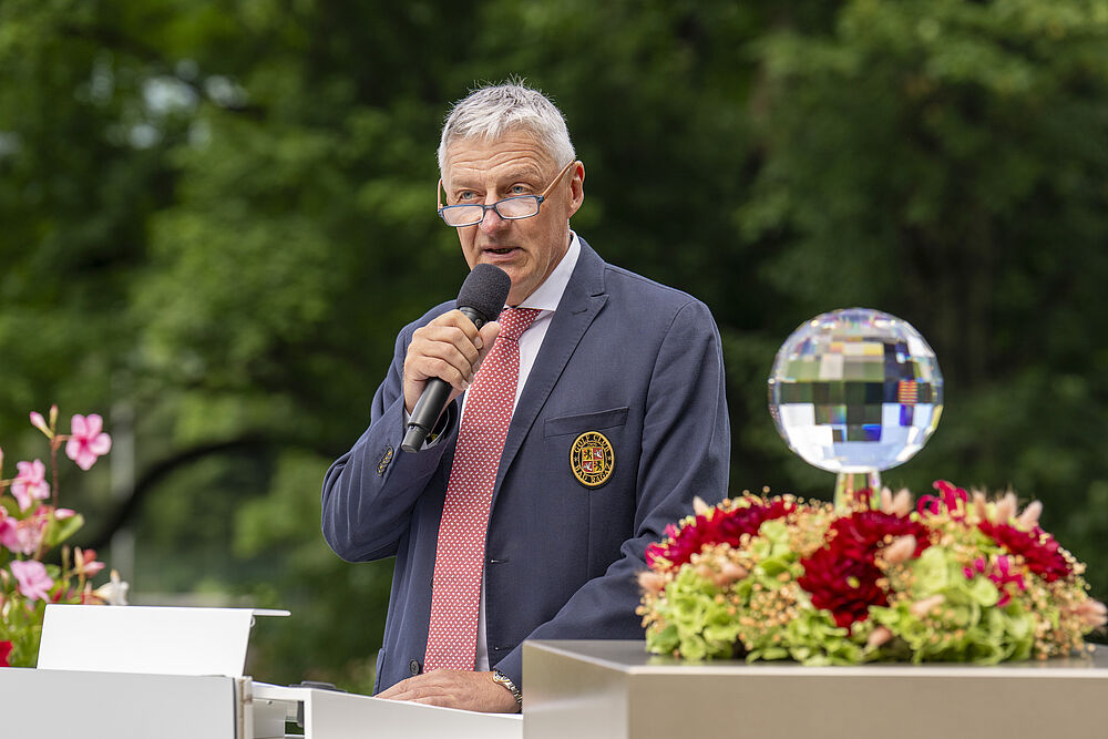 Eine Person im blauen Anzug und mit roter Krawatte spricht bei einer Veranstaltung im Freien in ein Mikrofon, das neben bunten Blumen und einer reflektierenden Kugel steht.