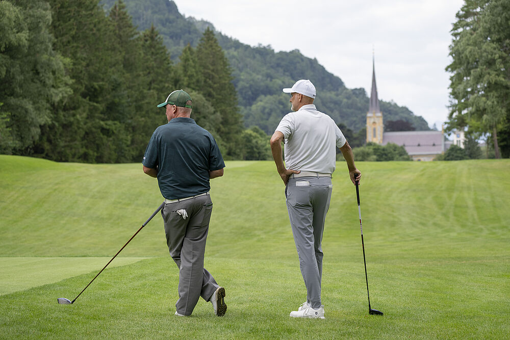 Zwei Golfer gehen mit ihren Schlägern in der Hand über einen grünen Platz. Im Hintergrund ist eine malerische Kirche zu sehen, eingebettet in baumbedeckte Hügel unter einem bewölkten Himmel.