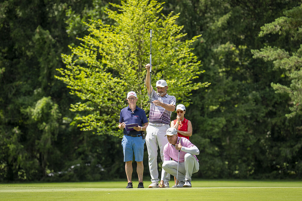Ein Golfer schwingt seinen Schläger auf einem üppig grünen Platz, beobachtet von zwei Personen. Das Sonnenlicht fällt durch die Bäume und schafft eine entspannte, konzentrierte Atmosphäre.