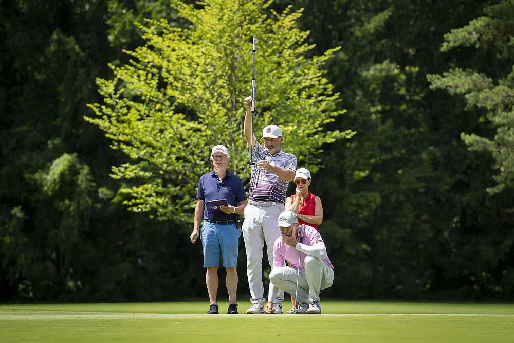 Ein Golfer, der mit dem Schläger die Linie sucht, wird von drei Personen auf dem Golfplatz beobachtet. Das helle Sonnenlicht wirft Schatten, und die Bäume bilden eine lebendige Kulisse.