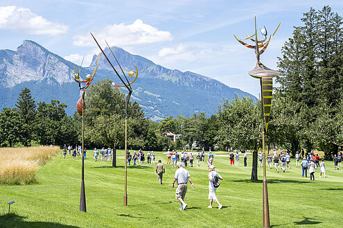 Die BadRagartz auf dem Golfplatz mit hohen, abstrakten Skulpturen, auf dem Menschen dem Turnier und Leaderflight folgen, vor einer Kulisse aus Bergen und Bäumen.