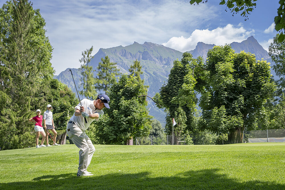 Ein Golfer beim Abschlag. Der Hintergrund mit viel Grün und Bäumen. Dahinter sind Berge zu sehen. Links neben dem Golfer stehen drei weitere Personen in rot und weiss gekleidet.