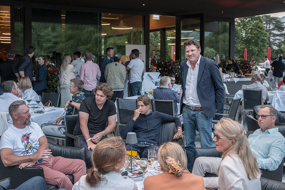 Eine Gruppe von Menschen an einer Party. Einer steht mit dunklem Jacket und lächelt in die Kamera. Die anderen sitzen und unterhalten sich.