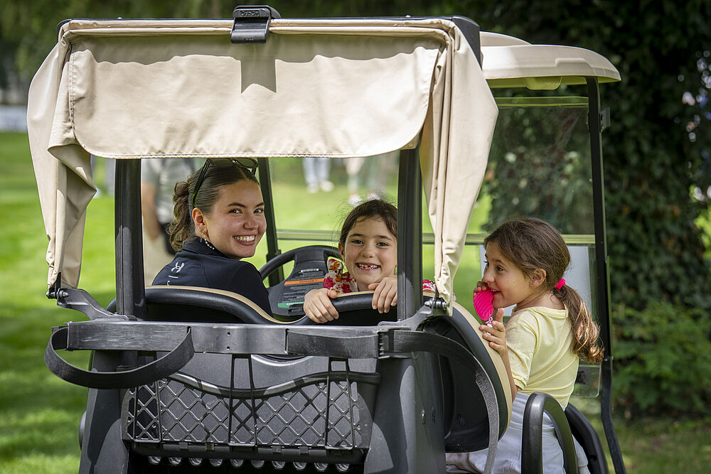 Eine Frau und zwei Kinder lächeln, während sie in einem Golfwagen fahren. Die Szene spielt sich in einem Park mit grünem Gras und Bäumen ab und vermittelt Freude und Freizeit.