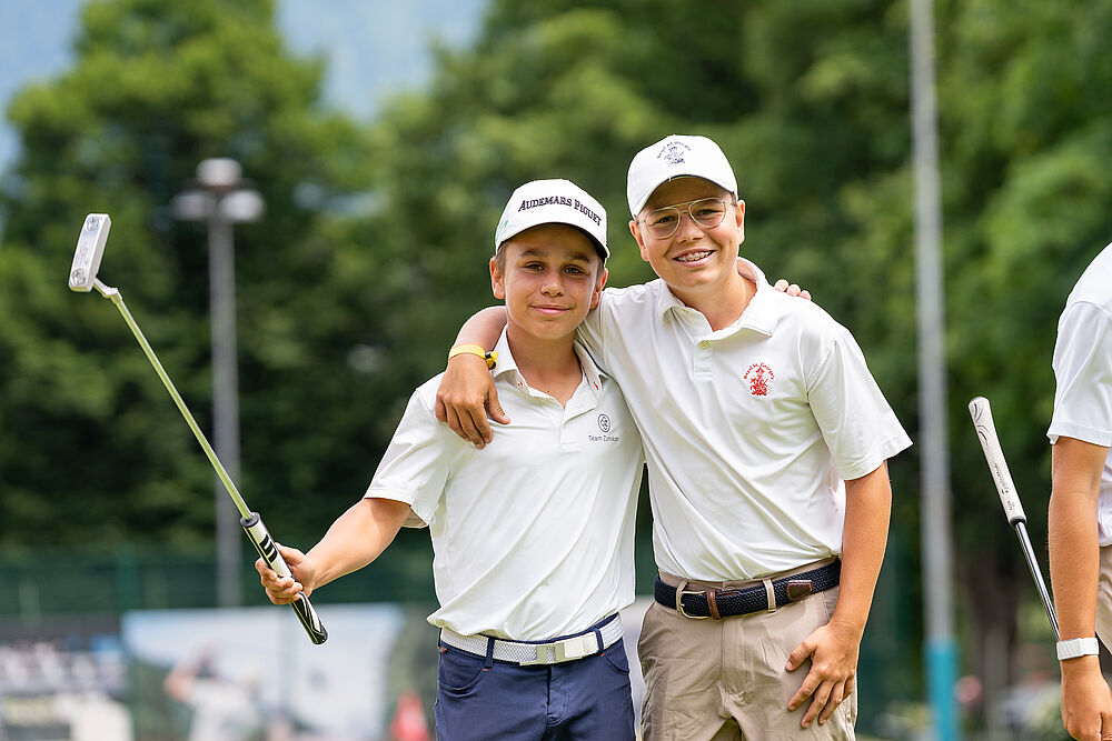 Zwei junge Golfer stehen nebeneinander und halten sich freundschaftlich. Beide tragen weisse Shirts und Caps. Einer hält einen Golfschläger in die Höhe. 