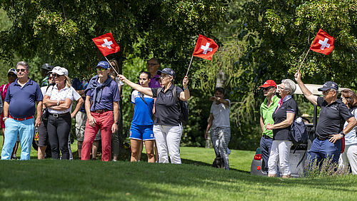 Eine Gruppe von Menschen steht auf einer Wiese und betrachtet die Pros auf dem Golfplatz. Zwei Helfer in schwarzen Tshirt schwenken enthusiastisch kleine Schweizer Fahnen. Die Szene ist lebendig, mit Bäumen im Hintergrund an einem sonnigen Tag.