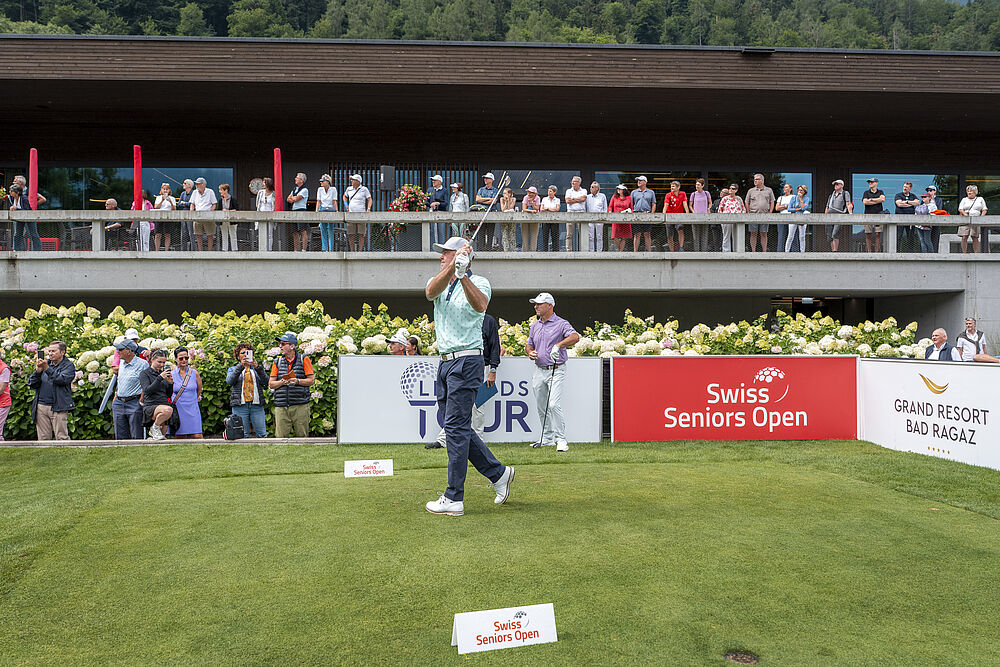 Mark Brown, der Sieger des Swiss Seniors Open 2025, schlägt ab zu seiner dritten und letzten Runde. Er trägt ein hellblause Shirt und eine dunkle Hose mit weissen Schuhen und einem weissen Cap. Hinter ihm sind Sponsoren Banden und auf der Terrasse des Clubhauses stehen eine menge Zuschauer.